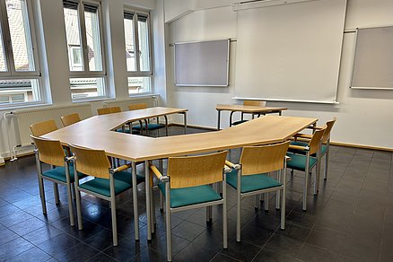 U-förmige Tischanordnung mit zehn Stühlen, an der offenen Stirnseite steht ein Einzeltisch mit einem Stuhl