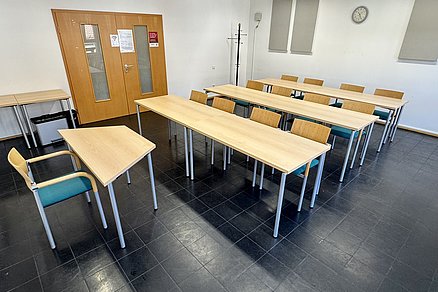 Drei Tischreihen zu je vier Stühlen blicken auf einen Tisch mit einem Stuhl an der Stirnseite