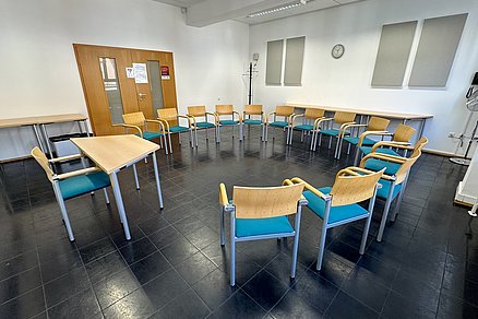 Ein offener Stuhlkreis mit vierzehn Stühlen blickt auf einen Tisch mit einem Stuhl an der Stirnseite