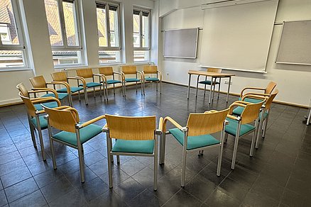Ein offener Stuhlkreis mit vierzehn Stühlen blickt auf einen Tisch mit einem Stuhl an der Stirnseite