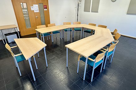 U-förmige Tischanordnung mit zehn Stühlen, an der offenen Stirnseite steht ein Einzeltisch mit einem Stuhl