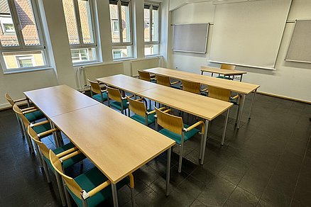 Drei Tischreihen zu je vier Stühlen blicken auf einen Tisch mit einem Stuhl an der Stirnseite